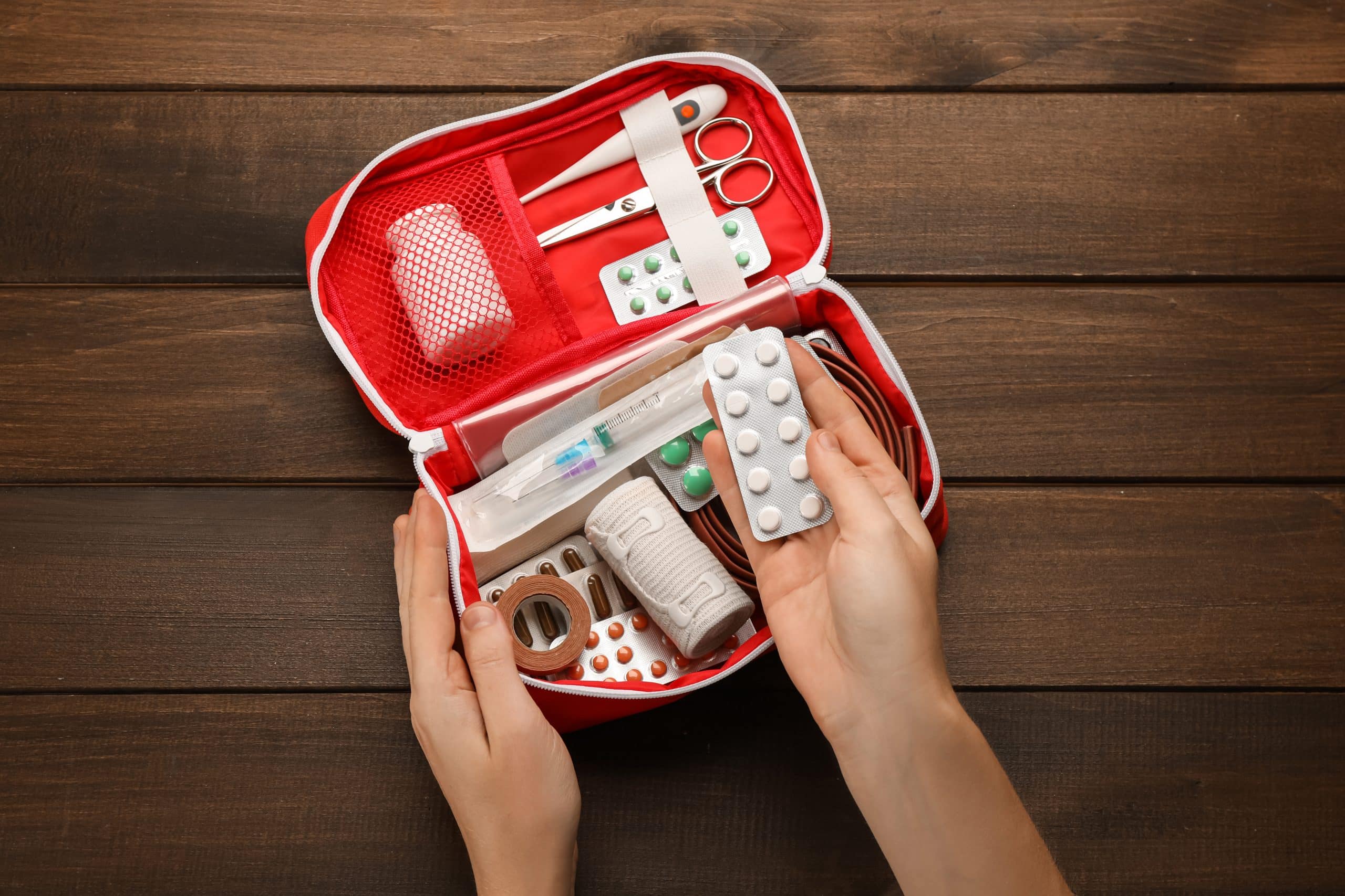 Geöffneter Erste-Hilfe-Kasten mit Medikamenten, Verbänden, Schere und Utensilien auf Holzoberfläche. Hand hält Blisterpackung mit Tabletten.